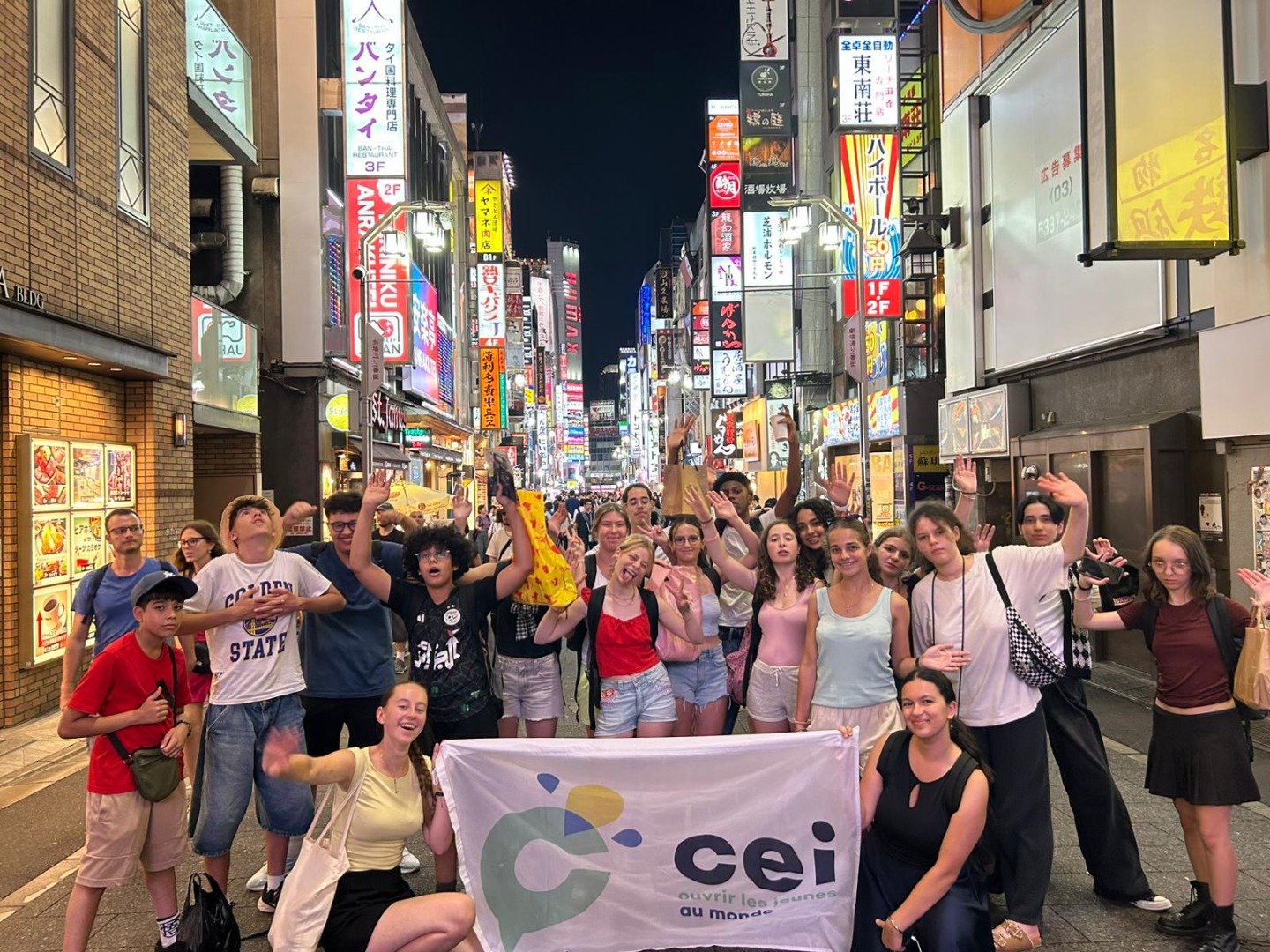 Groupe d’adolescents en colonie de vacances CEI à Tokyo, posant avec un drapeau CEI dans une rue animée et illuminée