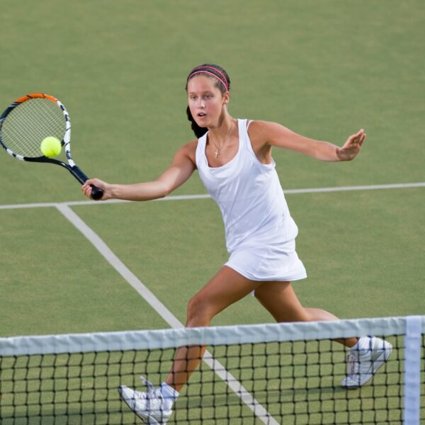 Une jeune joueuse de tennis en tenue blanche effectue une volée sur un court en gazon synthétique, illustrant la concentration et l'intensité d'un stage de tennis en immersion.