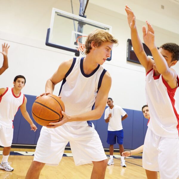 Un joueur de basketball en maillot blanc et bleu tient le ballon et cherche une opportunité de passe tandis qu'un défenseur en maillot blanc et rouge lève les bras pour bloquer son action. D'autres joueurs et un entraîneur sont visibles en arrière-plan dans un gymnase.
