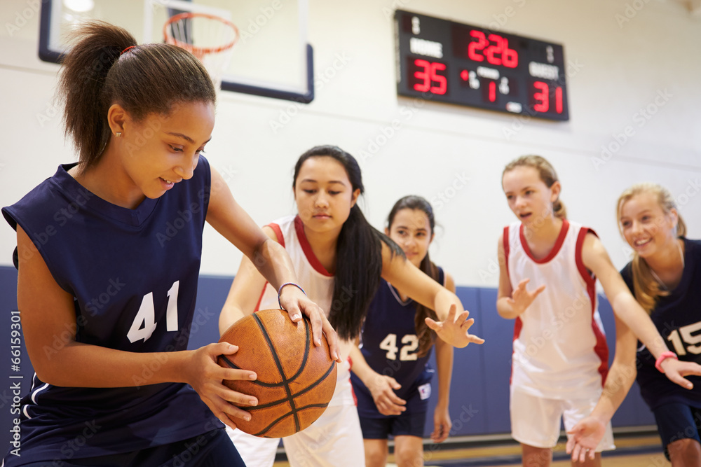 Une jeune joueuse de basketball en maillot bleu foncé protège le ballon sous la pression d’adversaires en maillot blanc et rouge, lors d’un match en salle. En arrière-plan, d’autres joueuses et un panneau d’affichage indiquant le score et le temps restant.