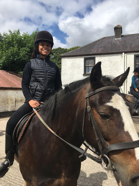 : Une jeune cavalière souriante, vêtue d'une tenue d'équitation noire et d'une bombe de protection, est assise sur un cheval bai dans une cour de ferme en Irlande, avec des bâtiments traditionnels et un autre cavalier en arrière-plan.